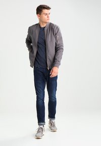 Giacca bomber grigia con zip frontale, polsini a costine e tasca sulla manica. Indossata sopra una camicia blu navy e abbinata a jeans scuri e sneaker.