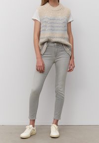 Chaleco de punto beige con rayas azules sobre una camiseta blanca, combinado con jeans skinny gris claro y zapatillas crema. Atuendo casual en capas.
