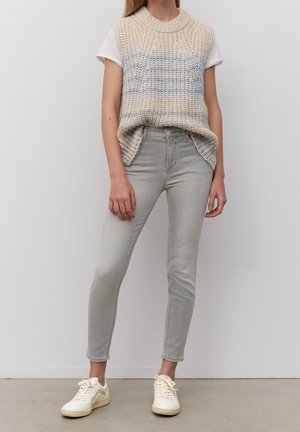 Chaleco de punto beige con rayas azules sobre una camiseta blanca, combinado con jeans skinny gris claro y zapatillas crema. Atuendo casual en capas.