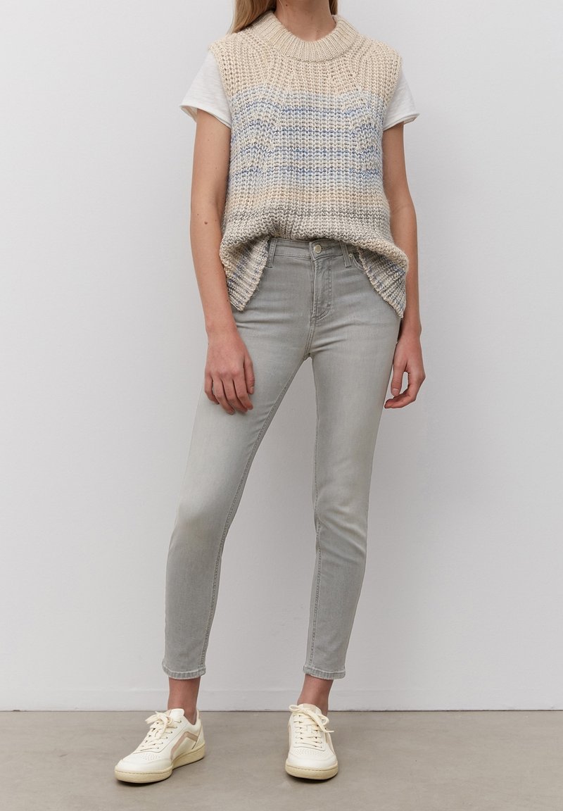 Chaleco de punto beige con rayas azules sobre una camiseta blanca, combinado con jeans skinny gris claro y zapatillas crema. Atuendo casual en capas.