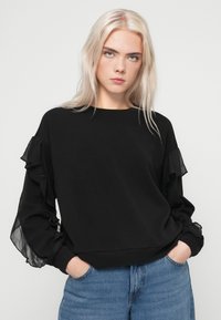 Czarny sweter z falbaniastymi rękawami z szyfonu, luźny krój, okrągły dekolt i prążkowane mankiety. Połączony z jasnoniebieskimi dżinsami.