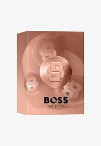 Caja de color rosa dorado con diseño redondeado, con logotipos en relieve y el texto "BOSS THE SCENT" en negro en la parte inferior, patrón minimalista.