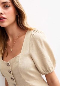 Beige linen dress with a square neckline, puff sleeves, and decorative buttons. Worn with a delicate gold necklace. Smooth texture.
