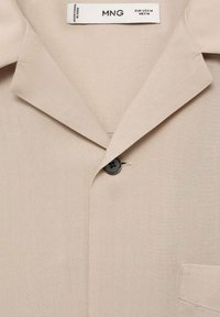 Beige, lichtgewicht stoffen overhemd met een uitgesneden kraag, een enkele zwarte knop en een zak aan de linkerkant. Schone, gladde textuur.