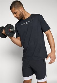 Schwarzes Baumwoll-T-Shirt mit kurzen Ärmeln, das ein weißes Logo auf der Brust trägt, kombiniert mit dunklen Shorts. Die Person hebt eine schwarze Kurzhantel.