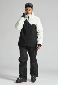 Chaqueta de esquí en blanco y negro, con un bolsillo con cremallera, capucha ajustable y tejido texturizado, combinada con pantalones de esquí negros y botas.