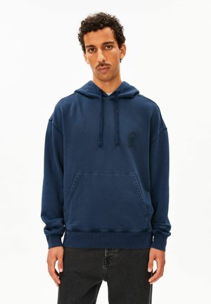 Homme aux cheveux bouclés et à la moustache, portant un sweat à capuche bleu marine avec poche avant et cordons, debout devant un fond blanc.