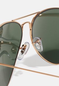Ochelari de soare tip aviator cu lentile verzi, ramă metalică aurie și suporturi pentru nas transparente. Dispune de un pod dublu și brațe subțiri.