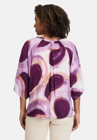 Lilla og creme bluse med et fluidt, abstrakt mønster, korte batwing ærmer, og en let plissé detalje bagpå. Glat stofstruktur.