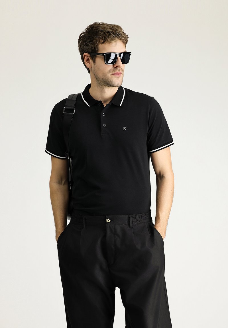 Polo nero con rifiniture bianche, maniche corte e patta con tre bottoni. Abbinato a pantaloni neri, accessoriato con occhiali da sole neri.