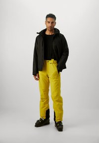 Black insulated jacket, black top, and bright yellow waterproof pants with black accents, paired with black boots for outdoor activity.