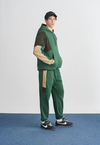Vihreä huppari ja siihen sopivat lenkkihousut, joissa on beigeä korostusta. Hupparissa on kengurutasku, ja mallilla on mustat kengät, joissa on valkoisia yksityiskohtia.