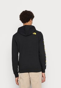 Schwarzer Pullover mit Kapuze, mit einem gelben Logo auf dem oberen Rücken und weißem Text entlang des Ärmels. Hergestellt aus weichem Stoff, lockere Passform.