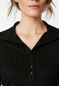 Schwarze gerippte Bluse mit weitem Kragen und Knopfleiste. Verfügt über sechs schwarze Knöpfe und eine dezente Textur über den Stoff.