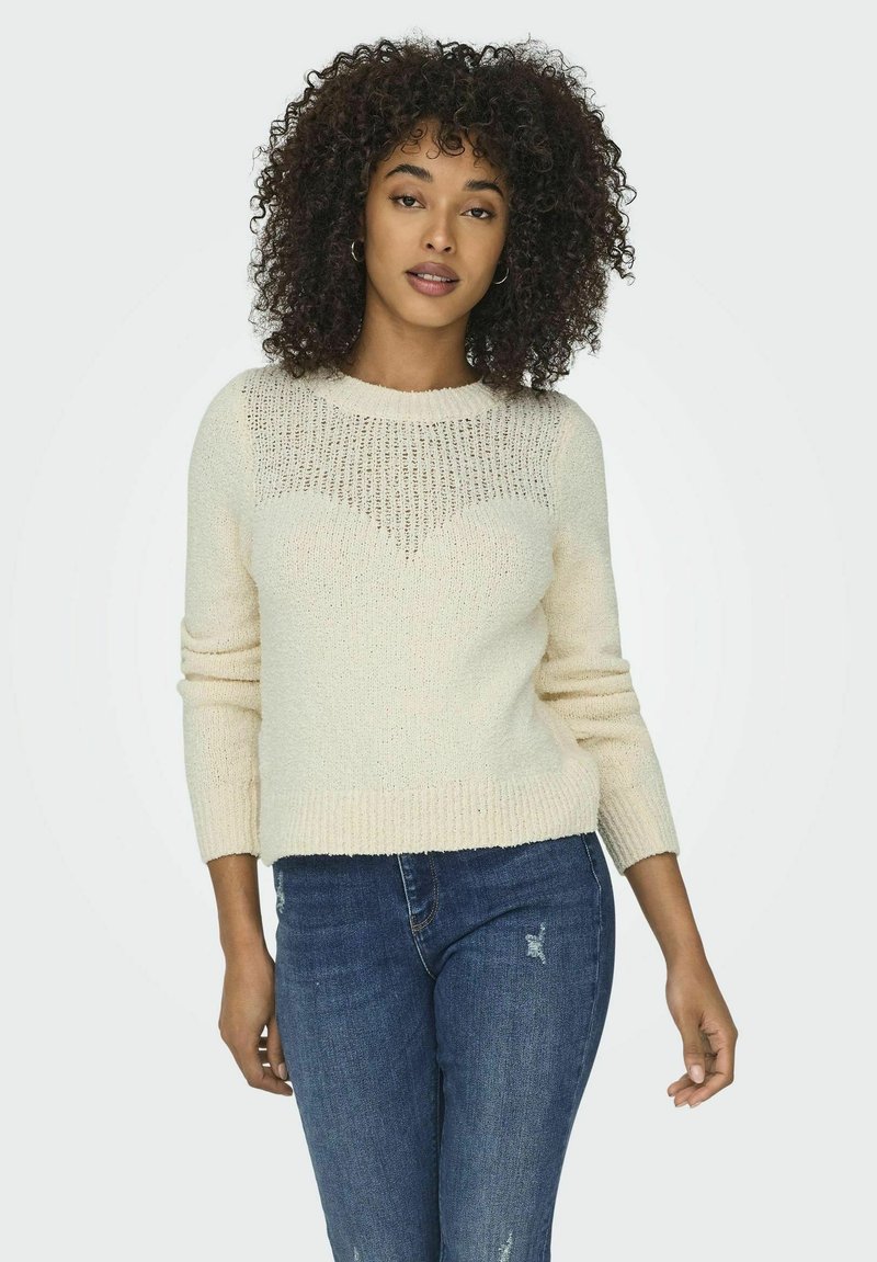 Pull crème à texture tricotée, avec un décolleté dégagé et des manches longues, associé à un jean bleu et à une coupe décontractée.