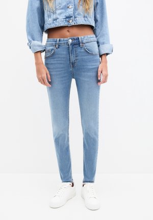Femme portant un jean skinny bleu clair, des baskets blanches et une veste en jean courte aux manches retroussées sur fond blanc.