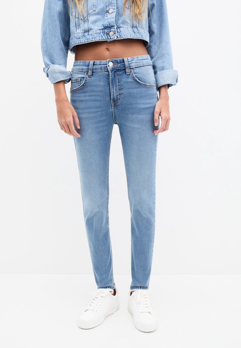 Femme portant un jean skinny bleu clair, des baskets blanches et une veste en jean courte aux manches retroussées sur fond blanc.