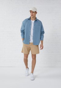 Helles blaues, gestreiftes Hemd über einem weißen T-Shirt, kombiniert mit khakifarbenen Shorts und weißen Sneakers. Akzentuiert mit einer beigen Kappe.
