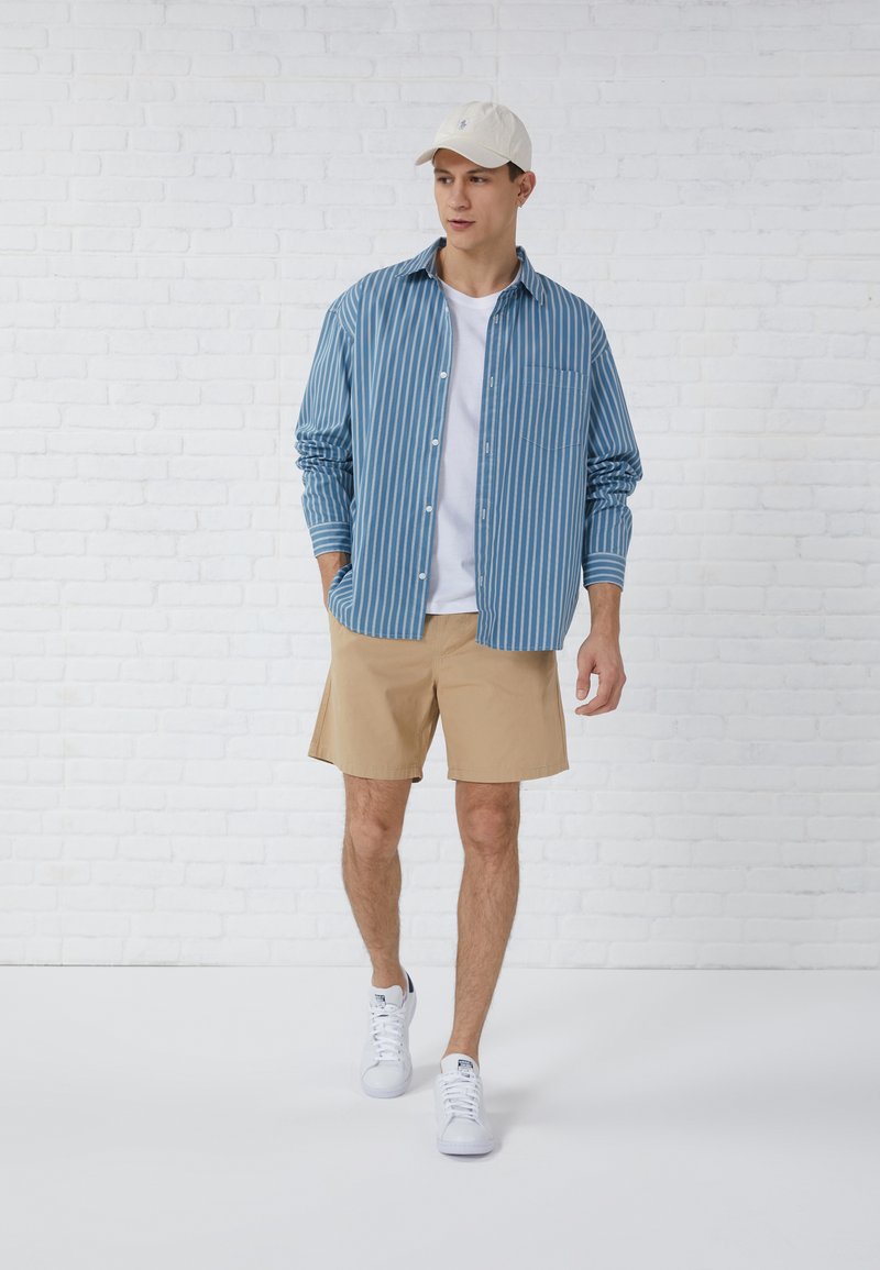 Helles blaues, gestreiftes Hemd über einem weißen T-Shirt, kombiniert mit khakifarbenen Shorts und weißen Sneakers. Akzentuiert mit einer beigen Kappe.