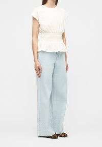 Femme debout portant un chemisier blanc à manches courtes avec taille smockée et un jean bleu clair à jambes larges avec des chaussures plates imprimé léopard.