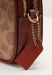 Sac en tissu marron avec un motif texturé, des garnitures dorées, des accents en cuir et une étiquette rouge embossée du mot « COACH », doté d'une fermeture éclair.
