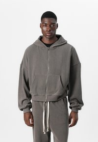Graue Zip-Hoodie aus strukturiertem Stoff mit lockerer Passform, großen Fronttaschen und einem Kordelzug am Saum. Kombiniert mit passenden Hosen.