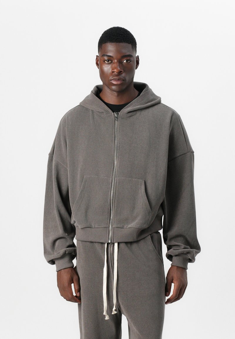 Graue Zip-Hoodie aus strukturiertem Stoff mit lockerer Passform, großen Fronttaschen und einem Kordelzug am Saum. Kombiniert mit passenden Hosen.