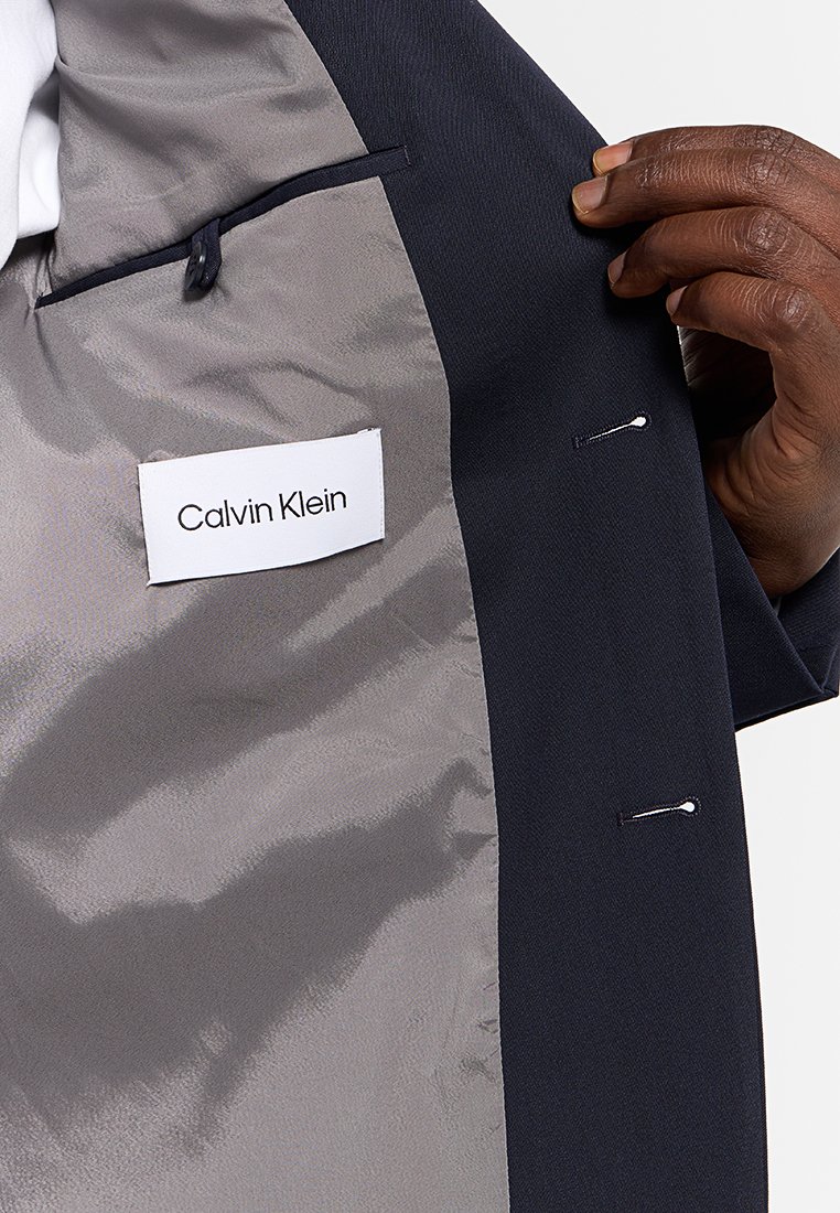 Intérieur d'un blazer bleu marine montrant une doublure grise lisse, une étiquette indiquant "Calvin Klein", deux poches cachées, et une main tenant la veste.