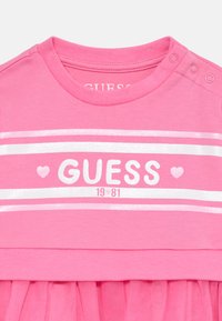 Pink cotton dress with short sleeves, featuring a rounded neckline, white stripes, and "GUESS 1981" text in metallic print. Tulle skirt detail.