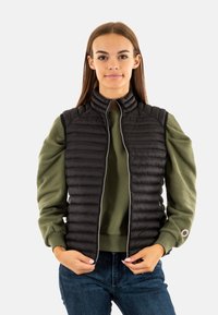 Chaleco acolchado negro con cuello alto, cierre de cremallera y patrón de acolchado horizontal, superpuesto sobre un sudadera verde. Unos vaqueros azules completan el atuendo.