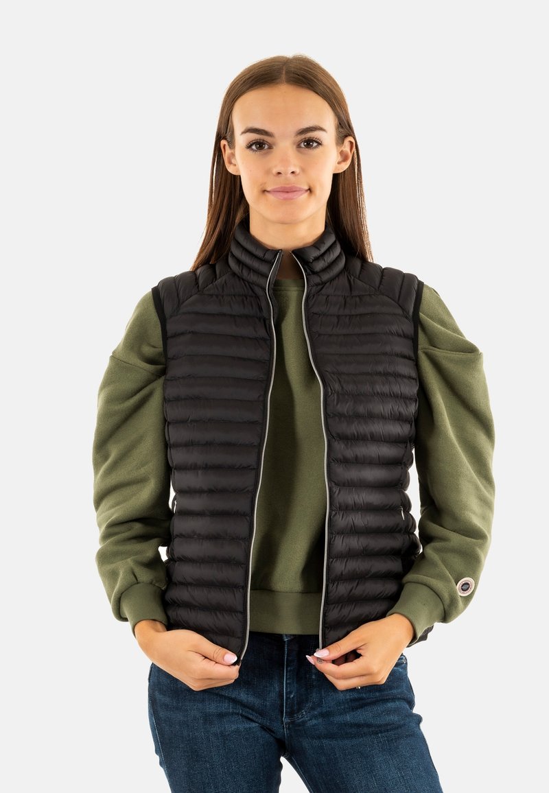 Chaleco acolchado negro con cuello alto, cierre de cremallera y patrón de acolchado horizontal, superpuesto sobre un sudadera verde. Unos vaqueros azules completan el atuendo.