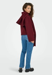 Bordeaux hættejakke med draperet rygdesign, parret med lyseblå lige ben jeans og sorte platform sko. Krøllet hår synligt.