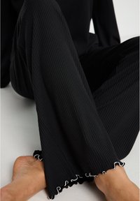 Pantalons en tissu côtelé noir avec une coupe décontractée ; présentent un ourlet froncé avec des coutures blanches. Vue rapprochée de la jambe et du pied.