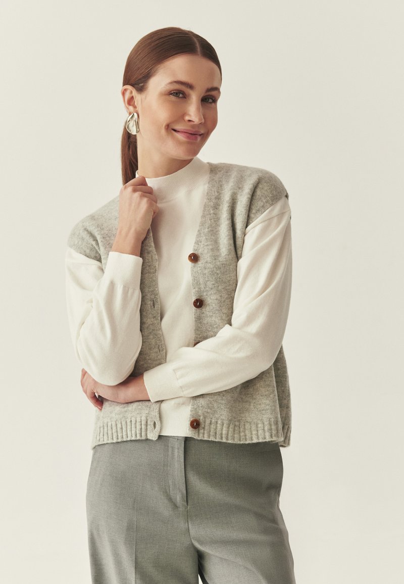 Light gray knitted vest with buttons, worn over a white long-sleeve top. The outfit is complemented by light gray trousers, featuring a ribbed hem.
