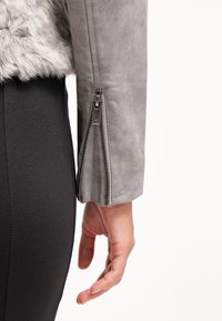 Main reposant à côté de la hanche, portant une veste en suède grise avec un détail de fermeture éclair sur la manche et un pantalon noir ajusté avec une couture visible.