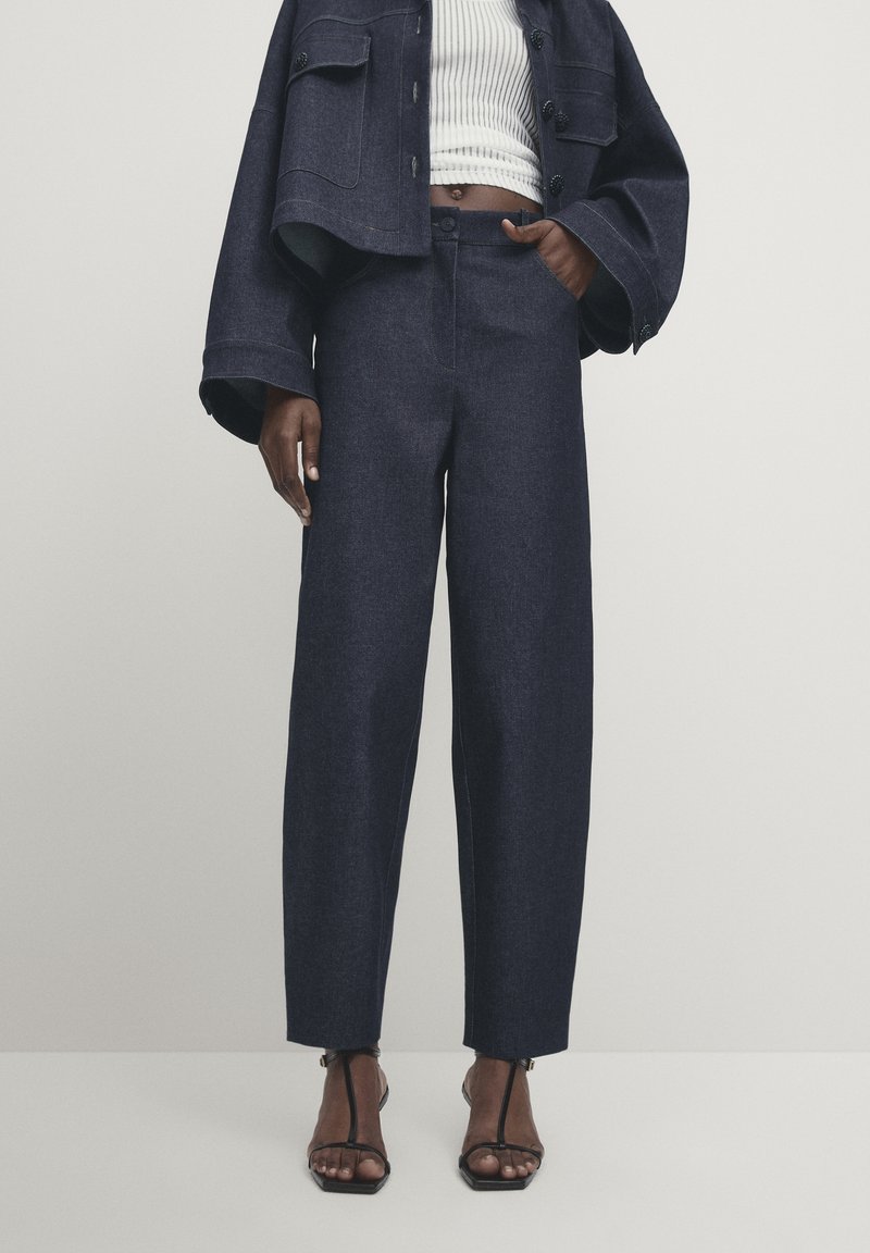 Un pantalon large en denim foncé avec une taille haute, des poches avant et une coupe structurée. Associé à une veste noire courte et des sandales à brides.