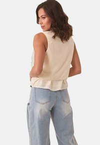 Top sin mangas de color beige claro con un dobladillo con volantes, con un respaldo texturizado y un patrón geométrico en verde claro. Combinado con jeans de mezclilla azul claro.