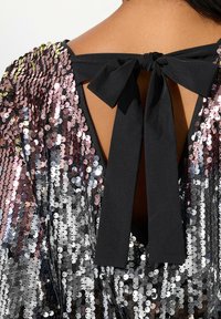 Top de lentejuelas con un lazo de cinta negra en la parte posterior. Presenta una mezcla de lentejuelas rosas y plateadas, creando una superficie texturada y reflectante.
