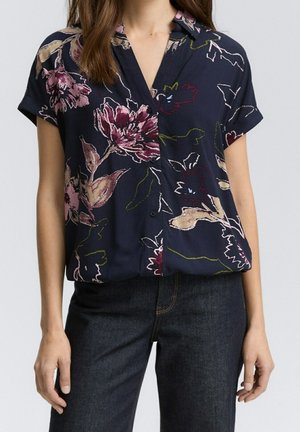 Kobieta ubrana w granatową koszulę z krótkim rękawem, zapinaną na guziki, z dużym różowym i bordowym kwiatowym wzorem oraz ciemnoniebieskie dżinsy.