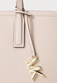 Beige leren totebag met een textuur, decoratieve gouden hardware charm en een smalle, dunne schouderband.