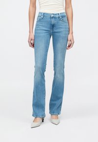 Femeie purtând blugi albastru deschis cu evazare, top alb cu pliuri și pantofi cu toc ascuțiți albi, stând pe un fundal alb simplu.