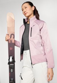 Giacca isolante rosa con accenti neri, vestibilità slim e tasche con zip, abbinata a pantaloni bianchi e a uno sci marrone con attacchi e grafiche.