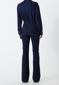 Jeans acampanados de denim azul oscuro con alta cintura, que cuentan con bolsillos traseros y una textura suave. El modelo lleva una camisa de denim a juego.