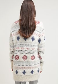 Chlupatý bílý kardigan s geometrickými vzory v červené, modré a námořnické barvě. Dlouhé rukávy, měkká textura a volný střih sahající pod boky.