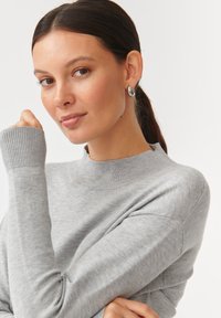 Sivi pulover s visokim izrezom i rebrastim manžetama. Mekana tekstura; prilagođen silueti. Model nosi jednostavne metalne naušnice.
