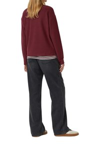 Vrouw staande, gekleed in een bordeauxrode lange mouwen top, gestreepte shirt daaronder, wijde donkere spijkerbroek en witte sneakers met bruine zolen.