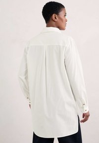 Biała koszula z długim rękawem z klasycznym kołnierzykiem, z detale w postaci plis na plecach oraz złotymi guzikami na mankietach, z prostym krojem i miękką teksturą.