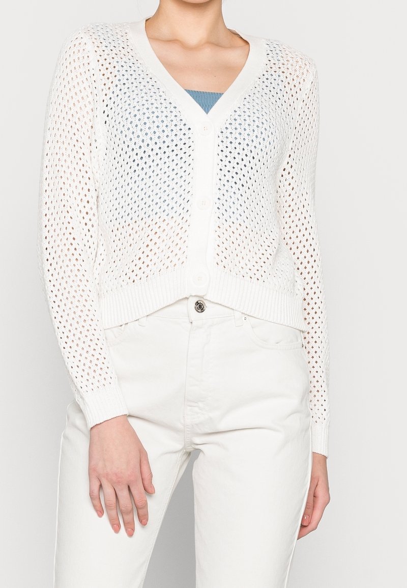 Cardigan blanc en tricot avec un col en V et une fermeture à boutons ; possède une trame ouverte et un modèle à mailles, ainsi que des poignets côtelés. Porté par-dessus un haut bleu clair.