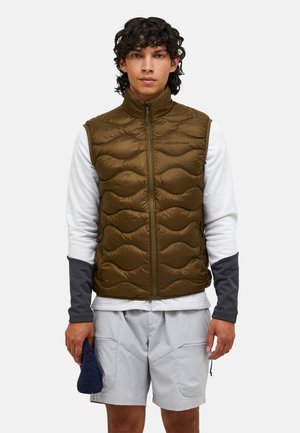 Giovane uomo che indossa un gilet trapuntato verde oliva sopra una camicia bianca a maniche lunghe e pantaloncini grigio chiaro, che tiene in una mano un cappello blu scuro.