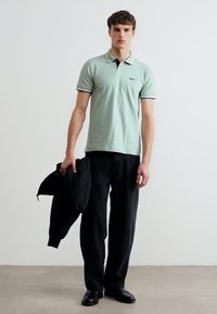 Νεαρός άνδρας στέκεται σε εσωτερικό χώρο φορώντας ανοιχτό πράσινο πόλο πουκάμισο, μαύρο παντελόνι, μαύρα παπούτσια, κρατώντας ένα μαύρο σακάκι στο αριστερό του χέρι.